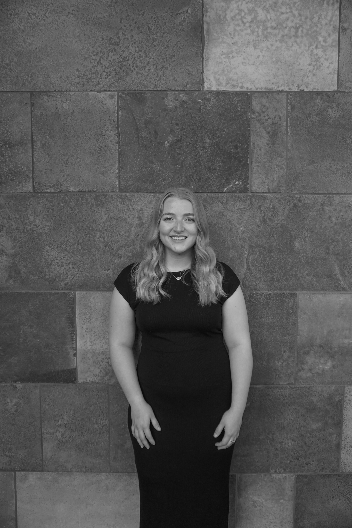 A person with long hair is smiling, dressed elegantly in a black dress, standing before a textured stone wall. It's the perfect look for a sophisticated Stafford dinner experience.