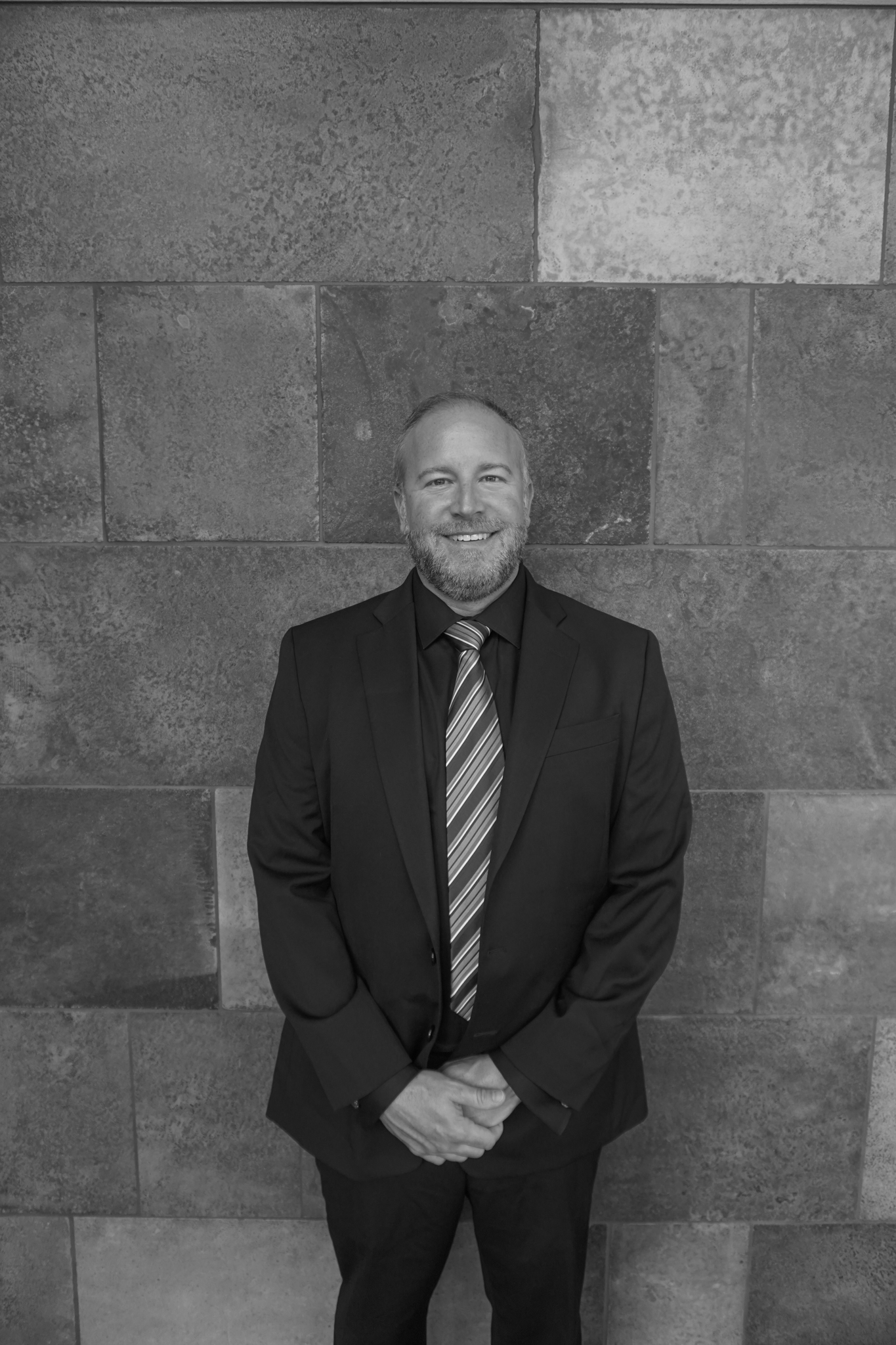 A man in a suit and tie stands smiling in front of a tiled wall, exuding the timeless elegance of a Stafford dinner. The image is captured in classic black and white, lending it an aura of sophistication.