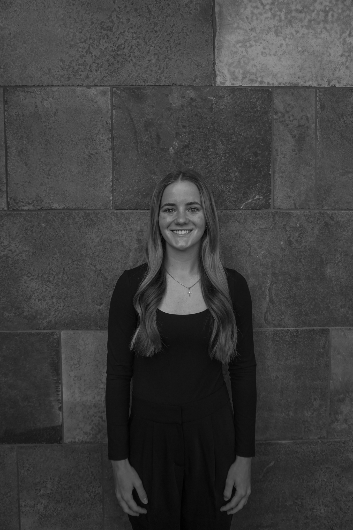 A person with long hair, wearing a dark outfit, stands smiling in front of a textured stone wall, seemingly having just left a delightful Stafford Dinner.