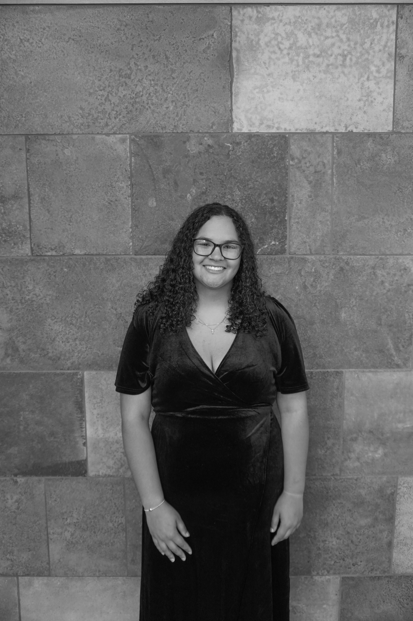 A person with curly hair and glasses stands smiling in front of a stone tile wall, elegantly dressed for a Stafford dinner in a dark wrap dress.