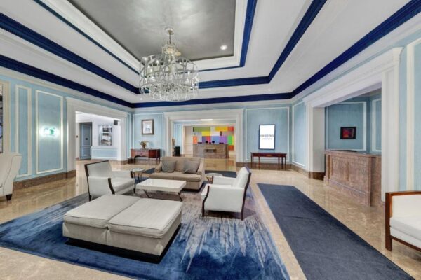 The lobby, adorned with light blue walls and a large chandelier, features white seating and a blue rug. A vibrant art piece near the reception adds flair to the elegant setting.