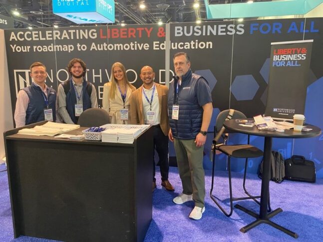 Five people standing at an automotive education booth with signs reading "Accelerating Liberty & Business For All.