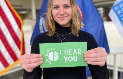 A person holds a green sign reading "I Hear You" with flags in the background, symbolizing Northwood University's commitment to mental health support.