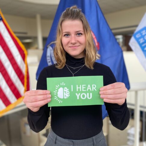 A person holds a green sign reading "I Hear You" with flags in the background, symbolizing Northwood University's commitment to mental health support.