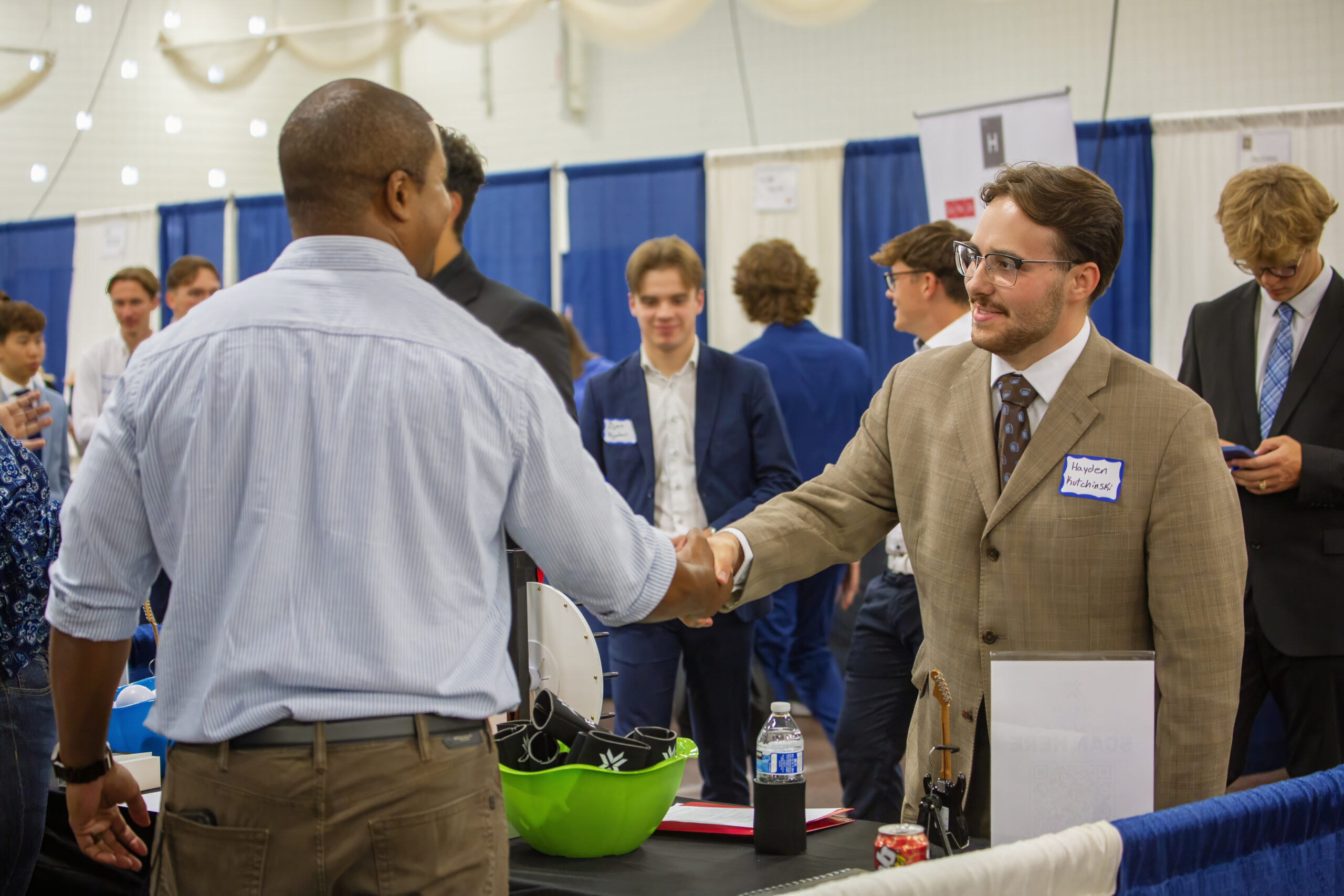 Two men dressed in business attire shake hands at a career fair. Tables with items and a water bottle are visible, along with other people in the background.