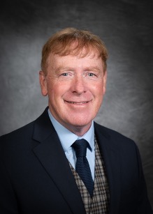 A person with short red hair wearing a suit, tie, and checkered vest, smiles against a gray background.