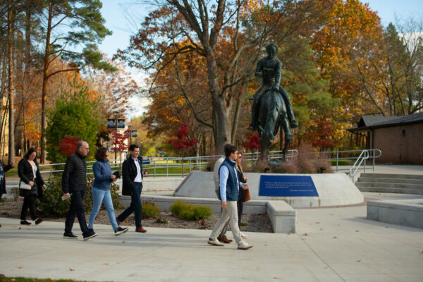 People walking past a statue of a person on a horse, surrounded by vibrant fall foliage, as they enjoy the scenic route that leads to Northwood University.