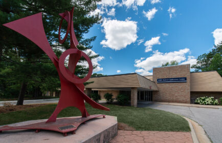 A modern red sculpture stands proudly in front of the NIU Naperville Conference Center, its vibrant hue contrasting with the brick facade. Framed by lush trees and a partly cloudy sky, this artistic landmark near the NADA Hotel reflects a harmonious blend of nature and creativity.