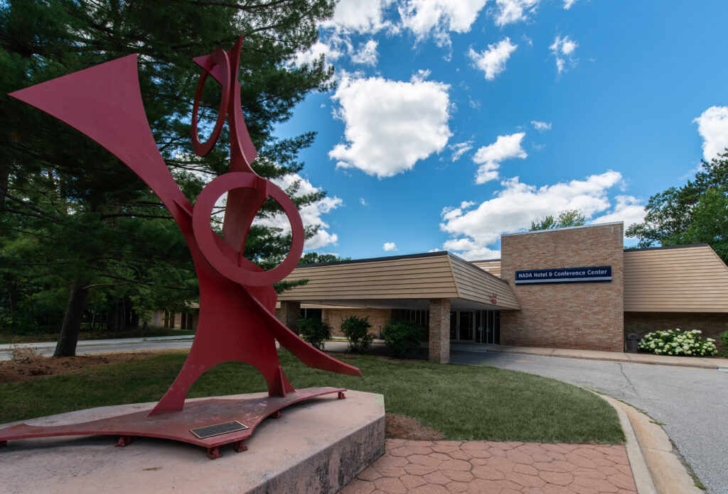 Red abstract sculpture in front of a brick building labeled "NASA Michoud Conference Center" with a cloudy sky and trees in the background.