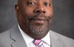 A bald man with a beard wears a gray suit and a pink-striped tie, posing against a dark gray background.