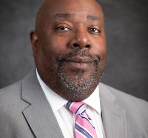 A bald man with a beard wears a gray suit and a pink-striped tie, posing against a dark gray background.