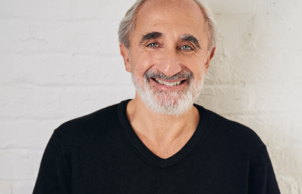 A man with gray hair and a beard smiles while wearing a black shirt, standing against a white brick wall.