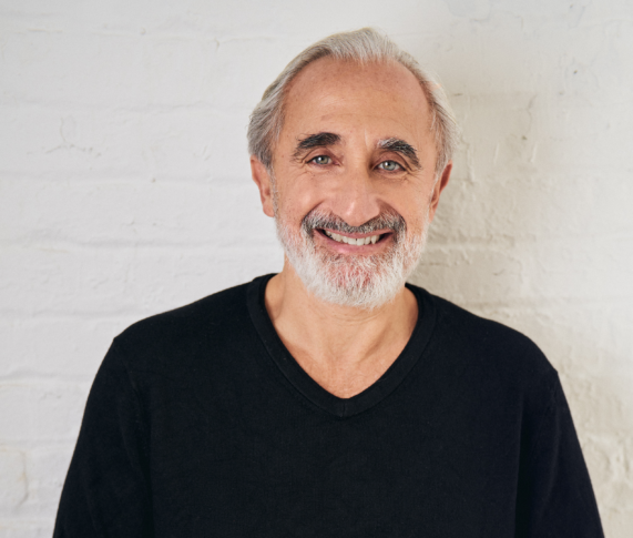 A man with gray hair and a beard smiles while wearing a black shirt, standing against a white brick wall.