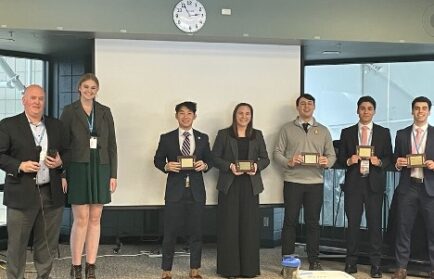 Seven people in formal attire stand in front of a screen, holding plaques. A clock shows 4:15. A projector is visible on the floor.