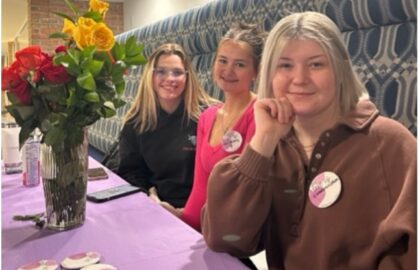 Three people sit at a table with a purple tablecloth, displaying buttons. A vase with red and yellow roses is on the table.