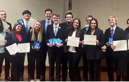 A group of fourteen people in formal attire stand posing with awards and certificates in a room. An American flag is visible in the background.