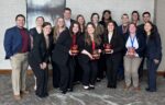 A group of fourteen people in business attire poses indoors, with some holding red awards.