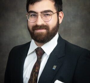 Person with dark hair, glasses, and a beard, wearing a black suit, white shirt, and patterned tie, in front of a gray backdrop.