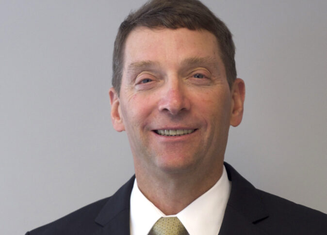 A man in a suit and tie stands against a plain background.