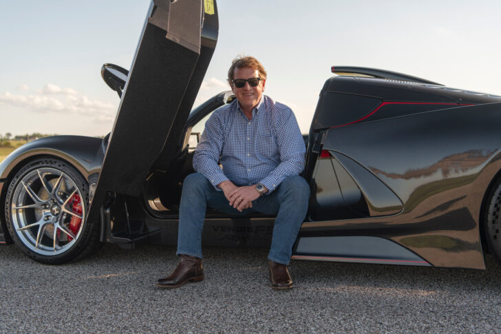 A man in sunglasses sits on the open door frame of a sleek black sports car parked on a road, with a clear sky in the background.