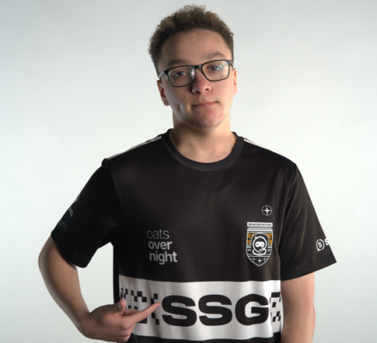 A person wearing glasses and a black esports jersey with white logos and text, including “oats overnight,” “SSG,” and “Northwood Esports,” points at the front of the shirt against a plain background.