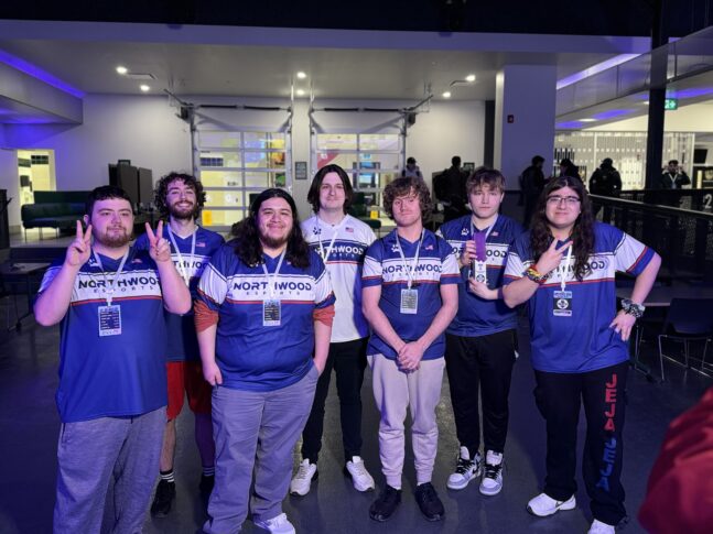 A group of eight people standing indoors, wearing matching blue and white jerseys with "Northwood" printed on them. Four are holding up peace signs.