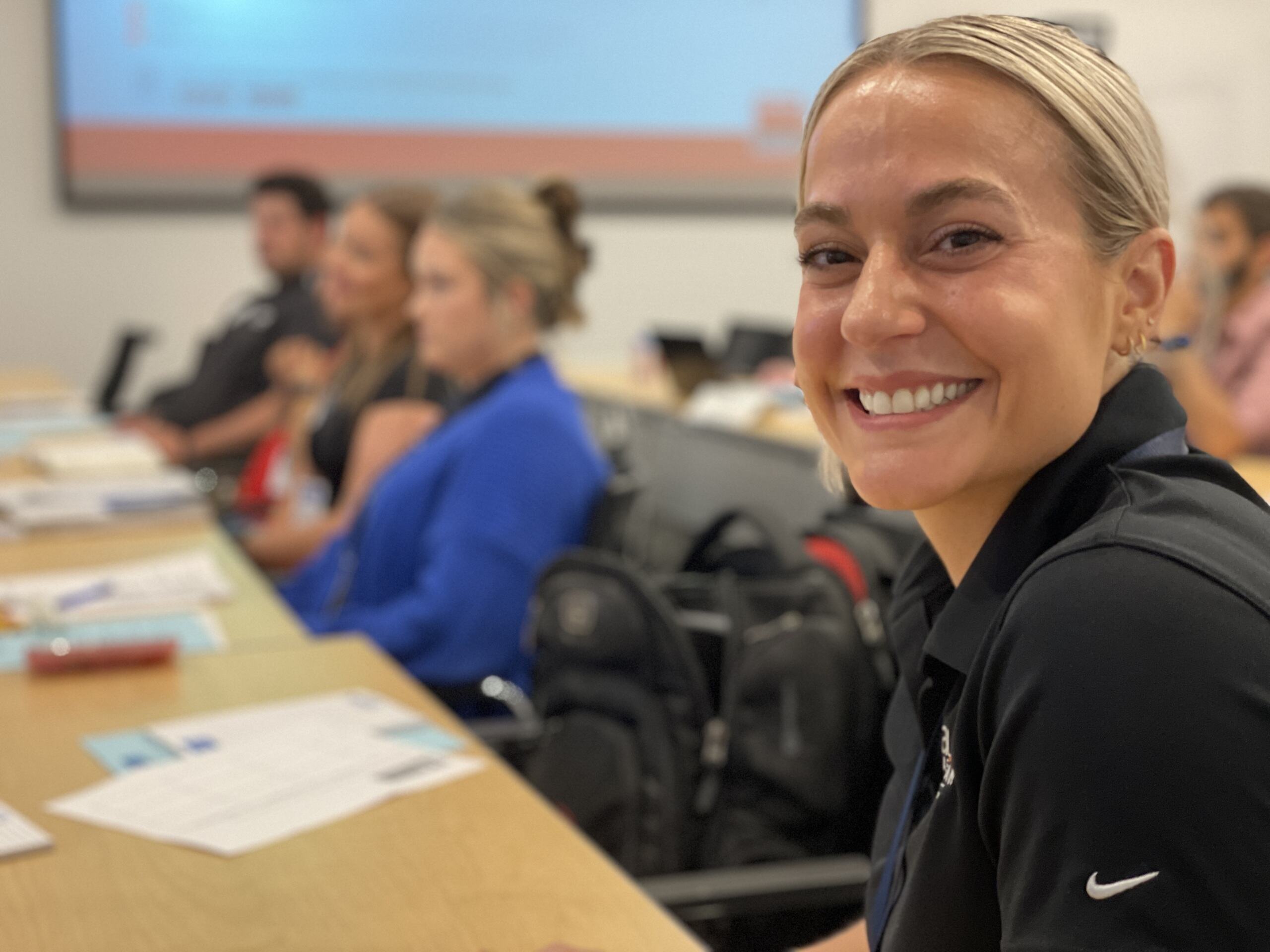Smiling person in focus at the front of a classroom setting with others blurred in the background.