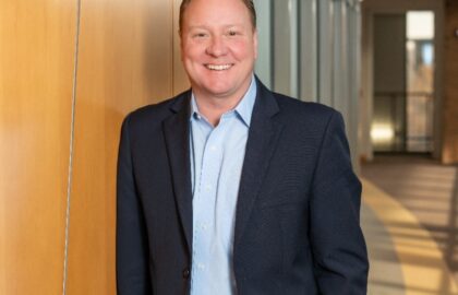 A man in a blue suit and light blue shirt stands smiling against a wood-paneled wall in a modern office hallway with large windows, featured in the Driven Newsletter, April 2025.