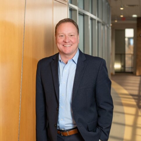 A man in a blue suit and light blue shirt stands smiling against a wood-paneled wall in a modern office hallway with large windows, featured in the Driven Newsletter, April 2025.