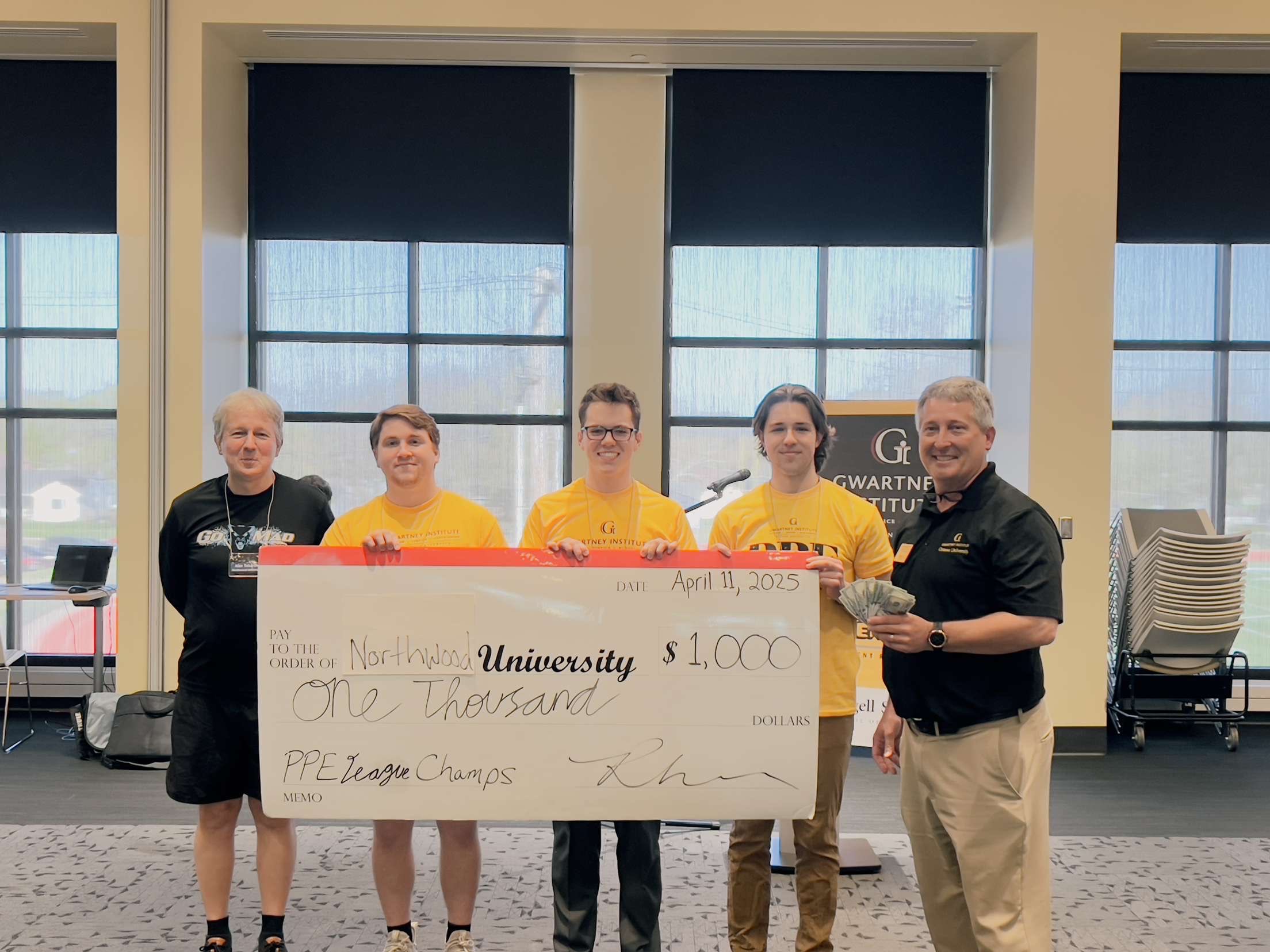 Five people stand indoors holding an oversized check for $1,000 made out to Northwest University for winning the PPE League Champs in Economics, Philosophy, and Politics, dated April 11, 2025.