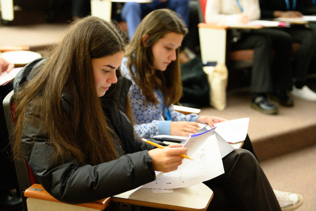 Two students sitting at desks in a classroom focus on writing and reviewing papers related to billing or payments during a class or exam.