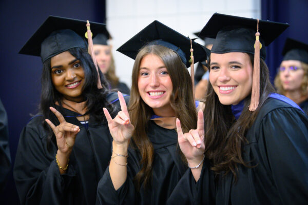 Three graduates in caps and gowns smile and make hand signs while posing for a photo at a graduation ceremony.