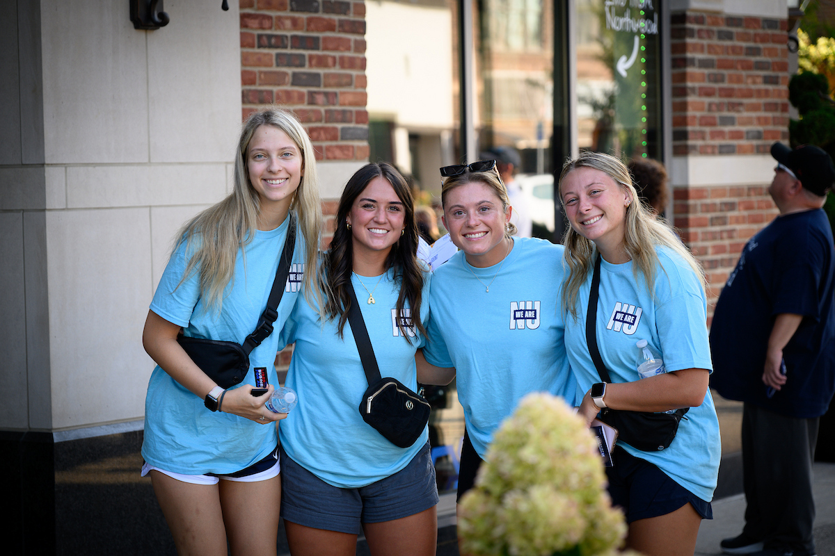 Four young women wearing matching light blue T-shirts stand together and smile outside a brick building at one of the universities in Midland, each carrying a small black bag.