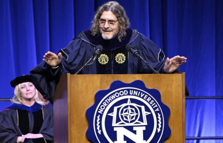 A man in academic regalia speaks at a podium with the Northwood University emblem, demonstrating leadership to the Class of 2025, while a woman in similar attire sits behind him on stage.