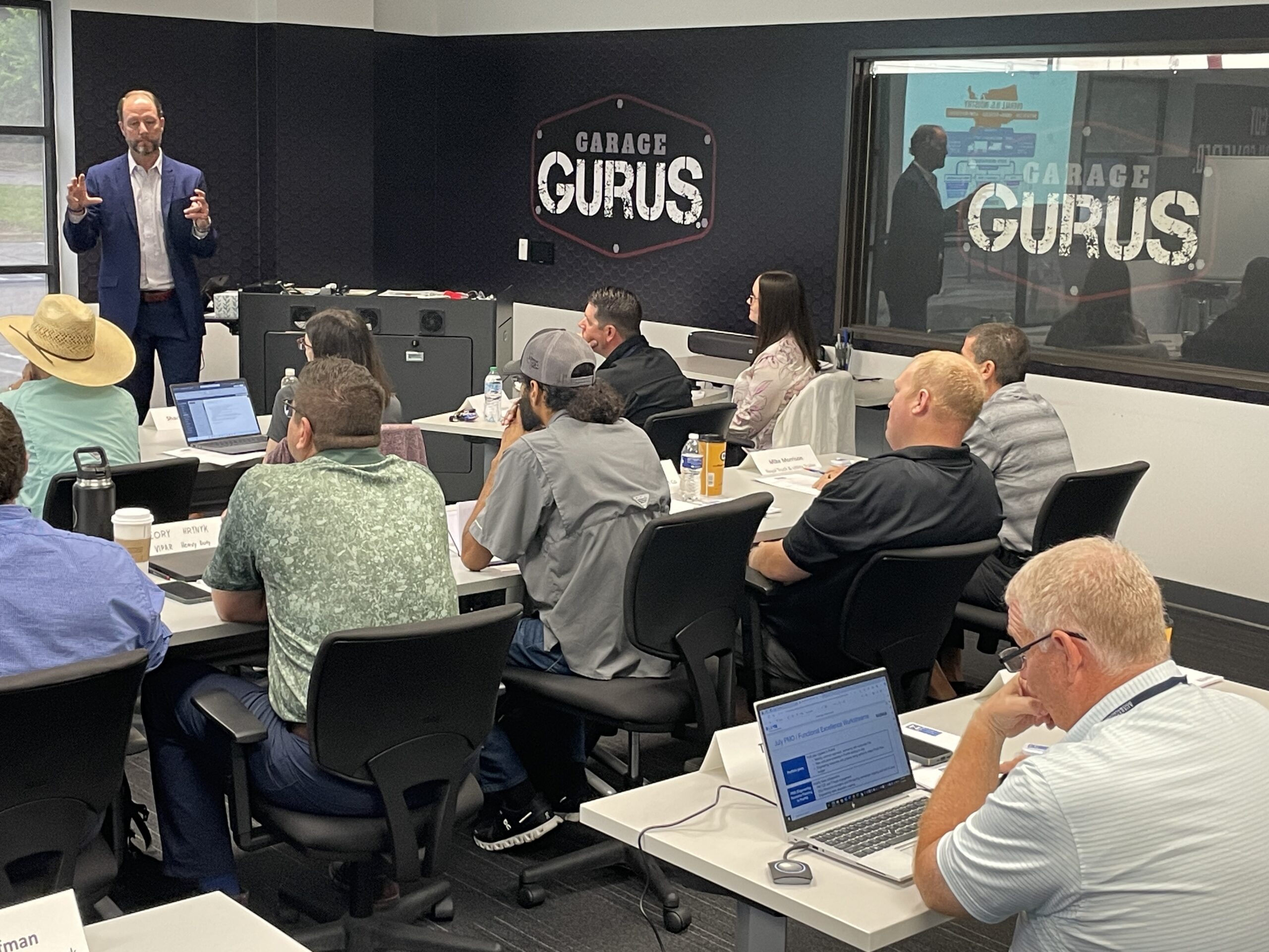 A man gives a presentation to a group of people seated at tables in a classroom labeled "Garage Gurus." Some attendees have laptops and notepads.