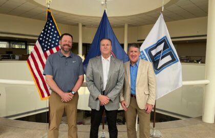 Three men stand indoors in front of the U.S. flag, another official flag, and a white flag with a logo, posing for a group photo in a spacious, well-lit setting.