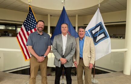 Three men stand indoors in front of the U.S. flag, another official flag, and a white flag with a logo, posing for a group photo in a spacious, well-lit setting.