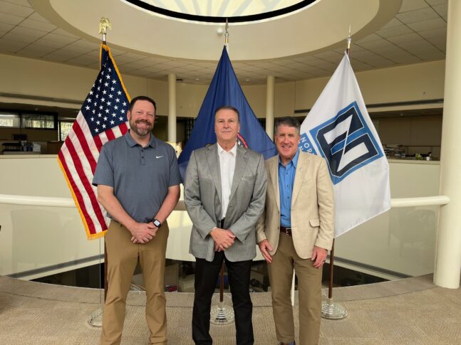 Three men stand indoors in front of the U.S. flag, another official flag, and a white flag with a logo, posing for a group photo in a spacious, well-lit setting.