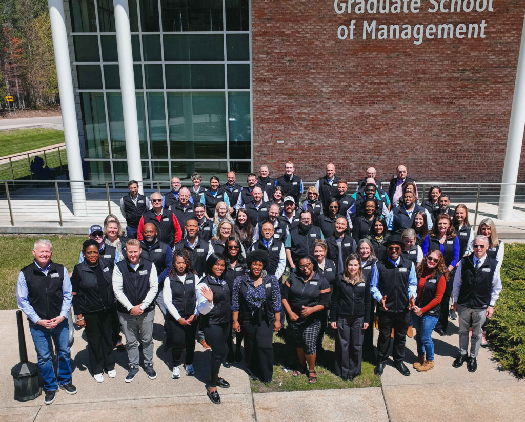 A large group of people, all wearing matching vests, stand outside a brick building labeled "Graduate School of Management," posing for a group photo on a sunny day.