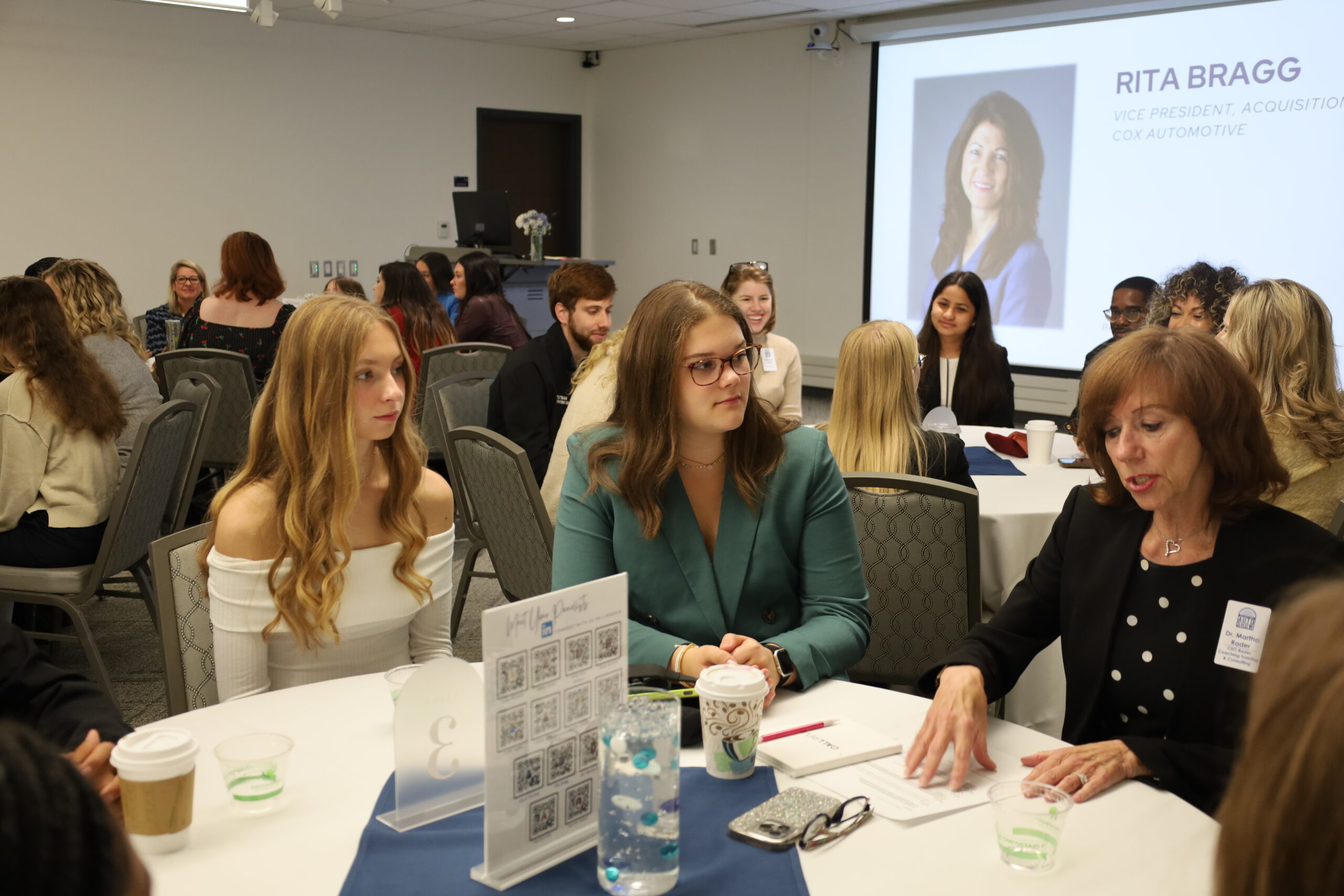 A group of people sit at round tables in a conference room, listening to a woman speak. A presentation slide behind them displays a photo and name: "Rita Bragg, Vice President, Cox Automotive.