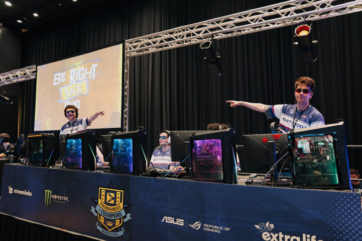 Esports players on stage at the Playfly Tournament of Champions in Arlington, Texas, point towards a screen displaying "Be Right Back," with gaming PCs and equipment visible in front of them.