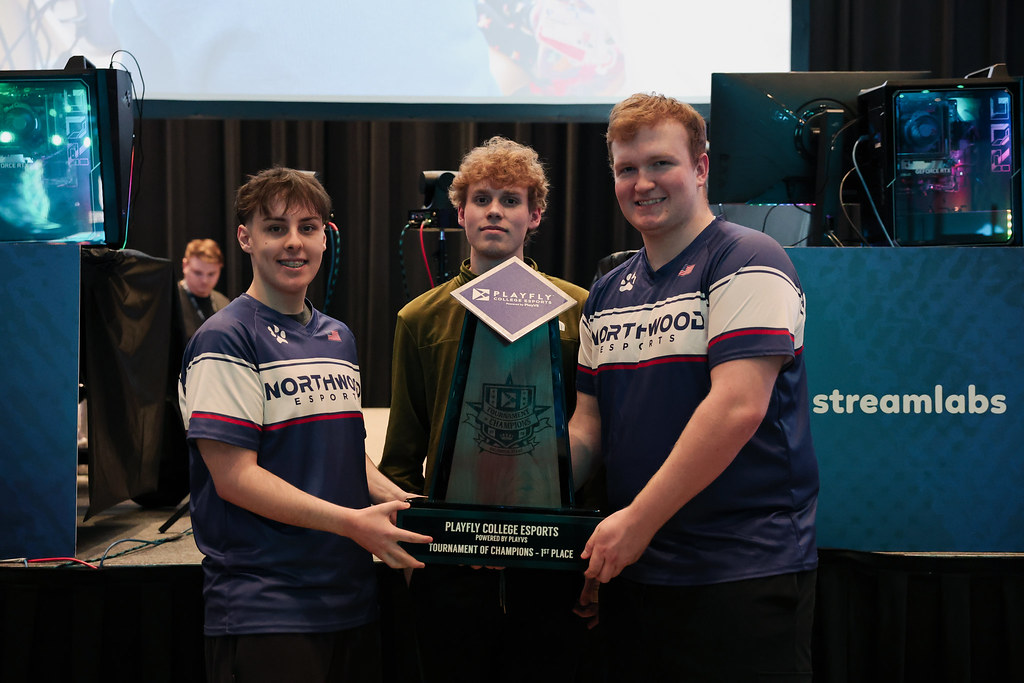 Three people stand indoors holding a large trophy labeled "Playfly College Esports Tournament of Champions—1st Place," with esports equipment and a streamlabs sign in the background.
