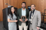 Three people stand indoors; the person in the center holds a plaque. All are dressed in business attire and smiling. A microphone and wooden paneling are visible in the background.