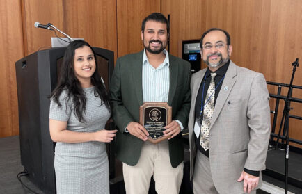 Three people stand indoors; the person in the center holds a plaque. All are dressed in business attire and smiling. A microphone and wooden paneling are visible in the background.