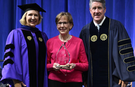 Three people in academic regalia stand together on stage; the woman in the center holds a glass award and smiles at the camera.