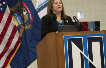A woman stands at a podium with an "N" logo, speaking in front of American and Michigan state flags during Northwood University’s Law Day.