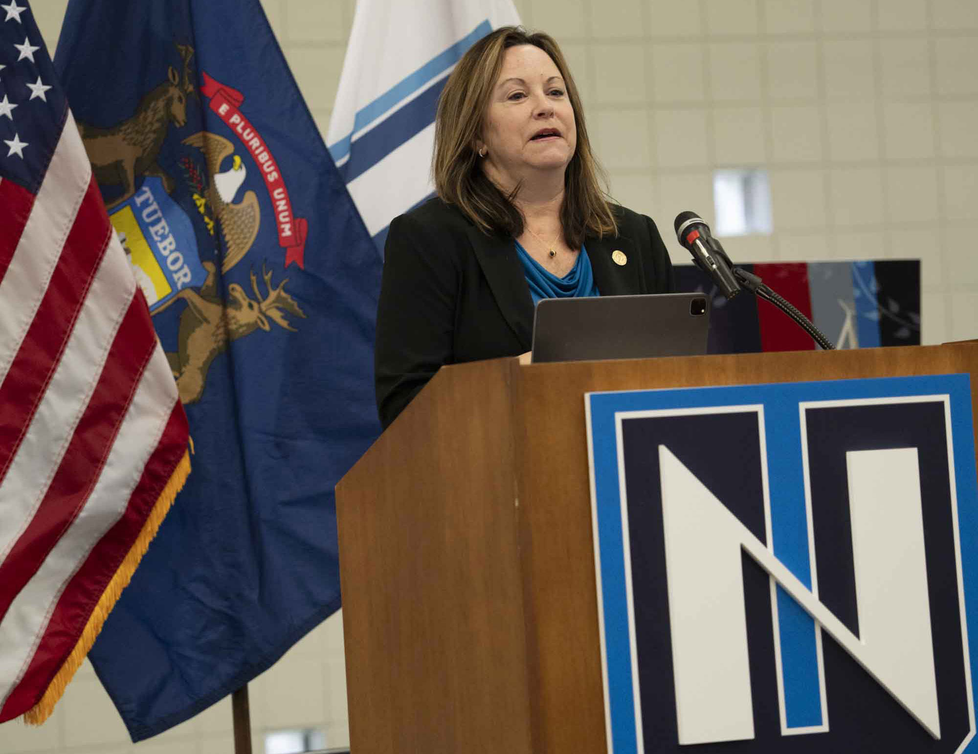 A woman stands at a podium with an "N" logo, speaking in front of American and Michigan state flags during Northwood University’s Law Day.