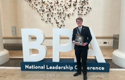A person in a suit stands holding a plaque in front of large "BPA" letters and a sign reading "National Leadership Conference" indoors.
