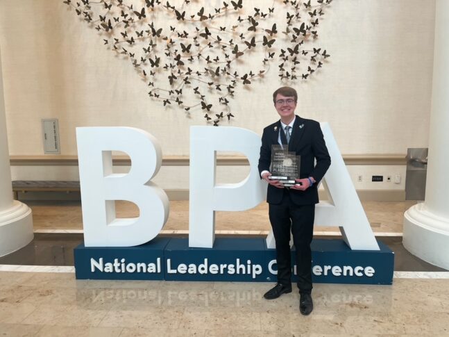 A person in a suit stands holding a plaque in front of large "BPA" letters and a sign reading "National Leadership Conference" indoors.