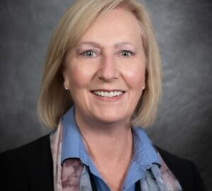 A woman with blonde hair smiles at the camera, wearing a blue collared shirt, patterned scarf, and dark jacket against a gray studio background.